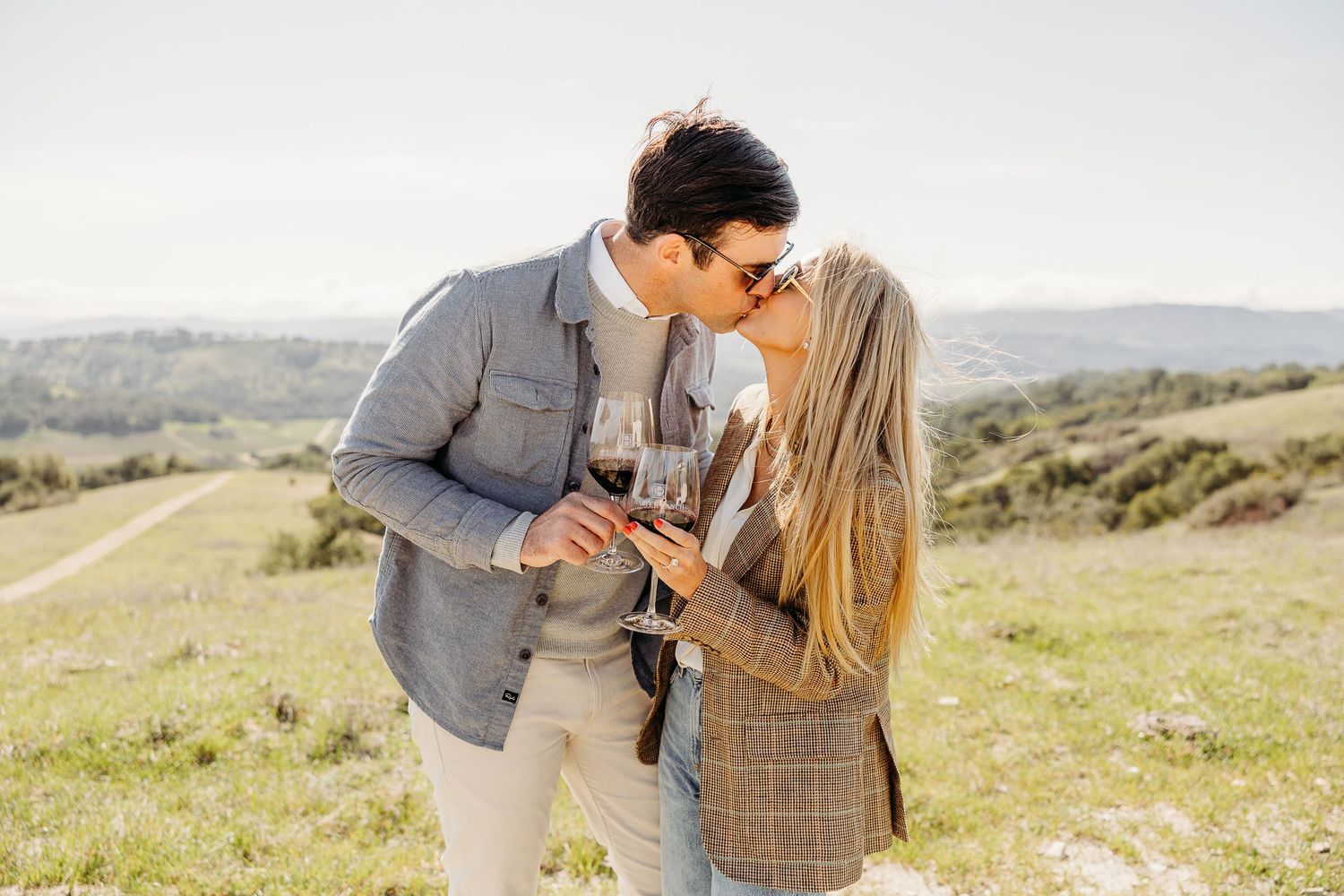 Couple embracing in a sun-lit meadow with rolling hills in the background.