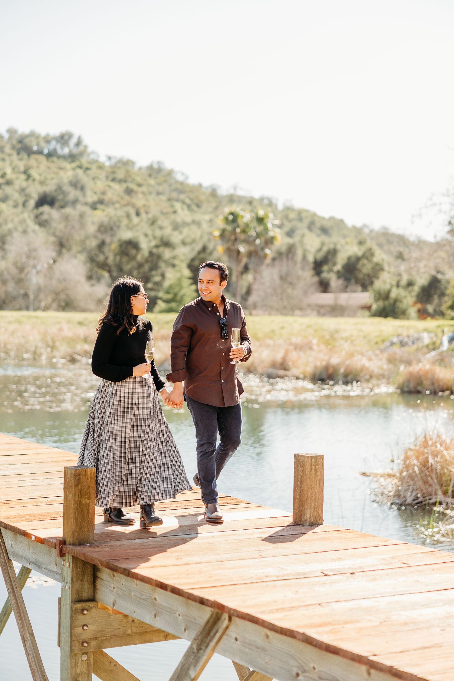 A couple enjoys a romantic moment holding hands while standing on a wooden dock by a serene lake.