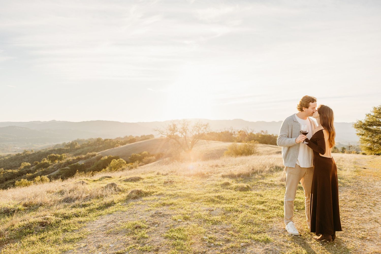 Silhouettes of a couple standing close together against a bright sunlit hillside landscape.