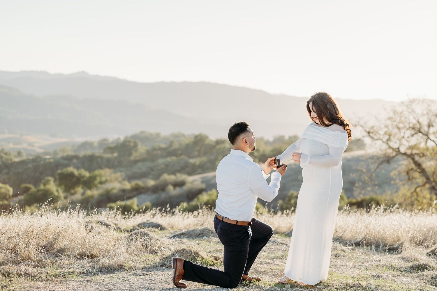 Man proposes to woman on one knee at golden hour with scenic mountain valley backdrop.
