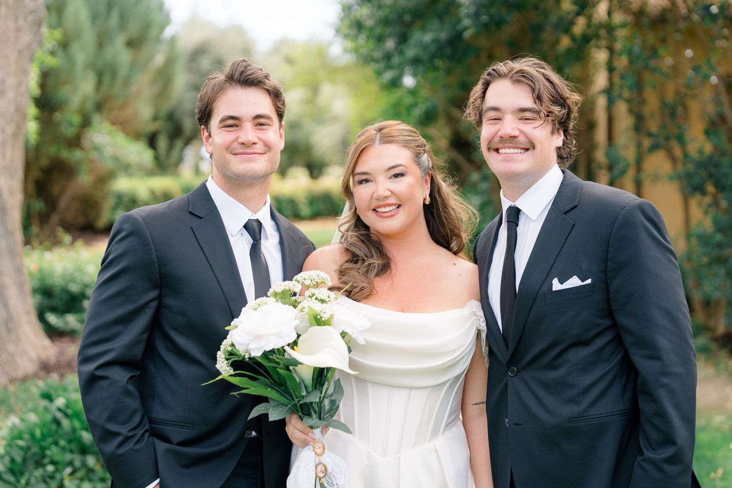 Bride in off-shoulder white gown poses with two groomsmen in black suits holding white bouquet outdoors.