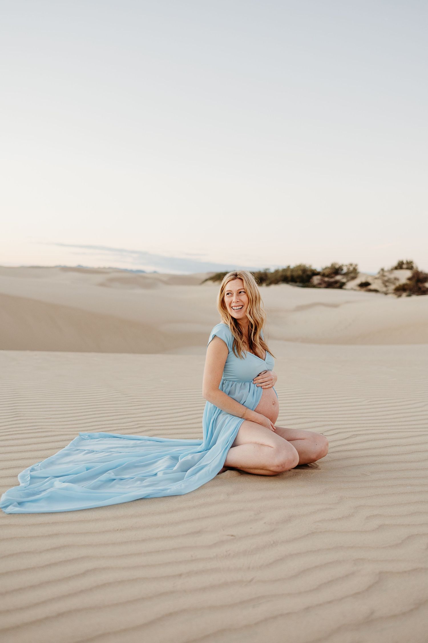 Serene desert maternity portrait with long blue dress trailing on sand.