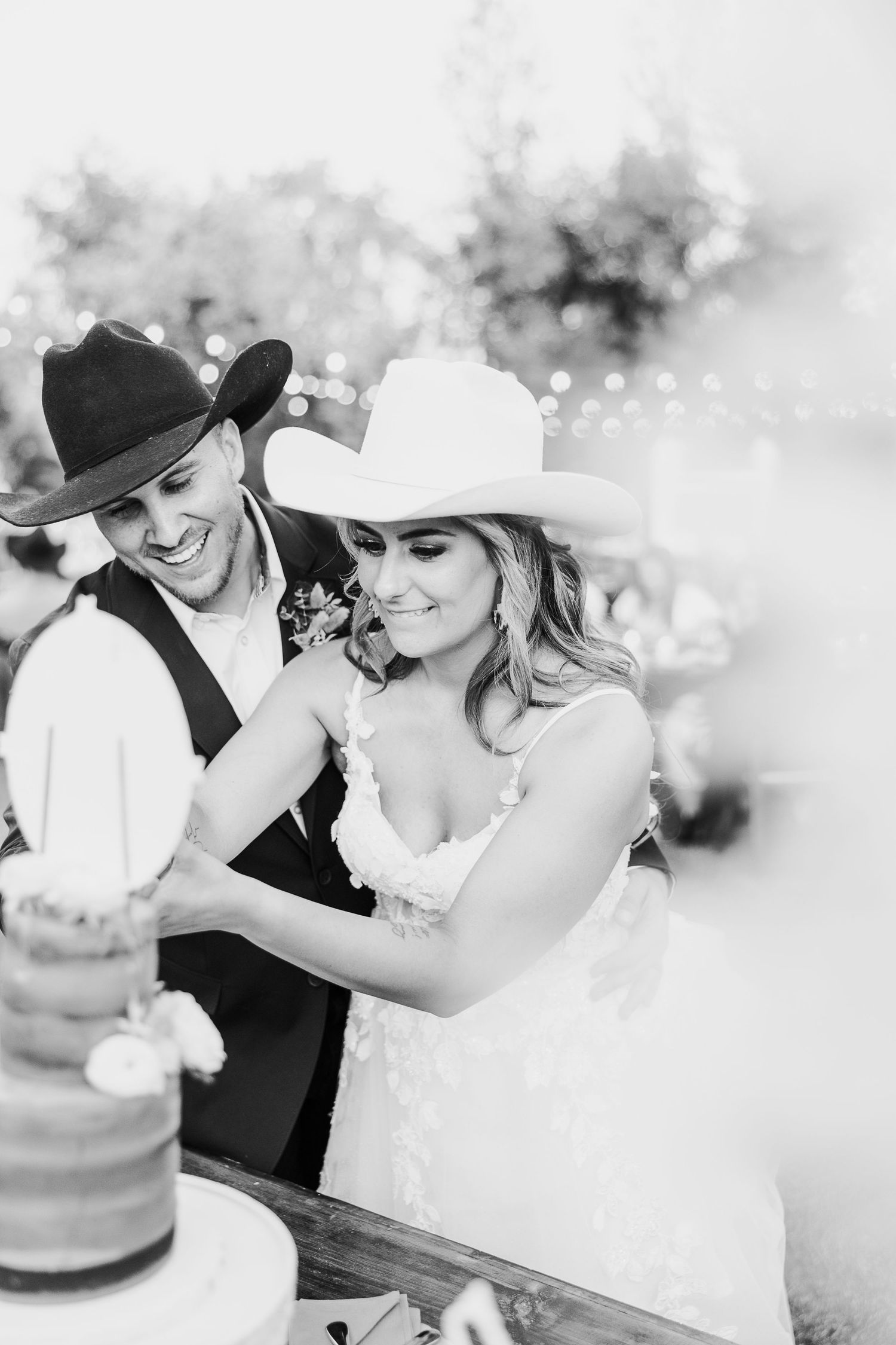 A couple in western attire cutting their wedding cake together at a rustic outdoor celebration.