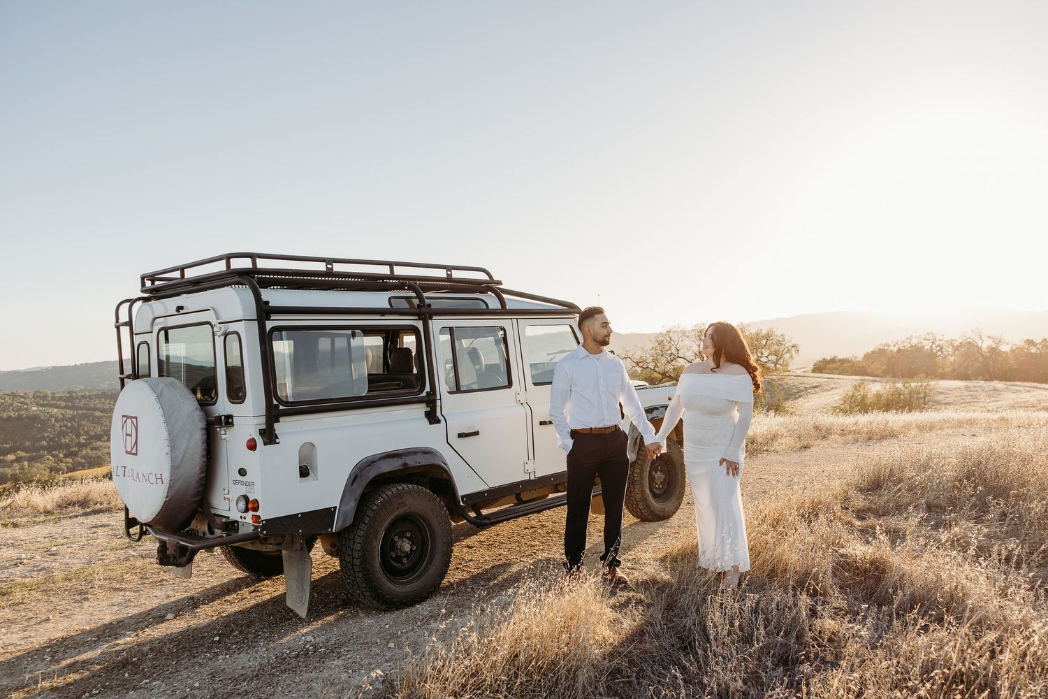 A white vintage Land Rover Defender parked in a golden field during sunset with people standing nearby.