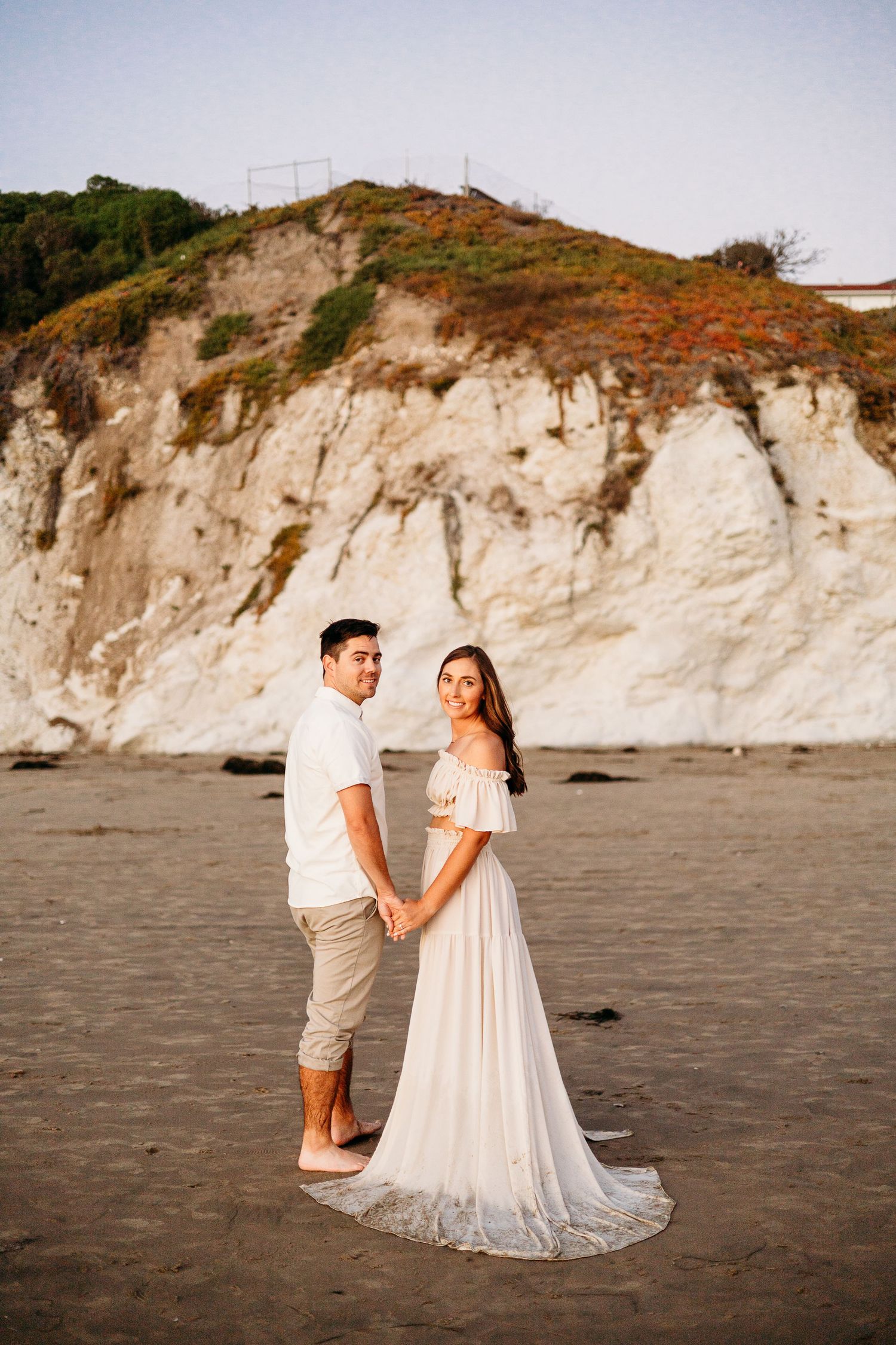 A couple holds hands on a beach with towering white cliffs behind them during golden hour.