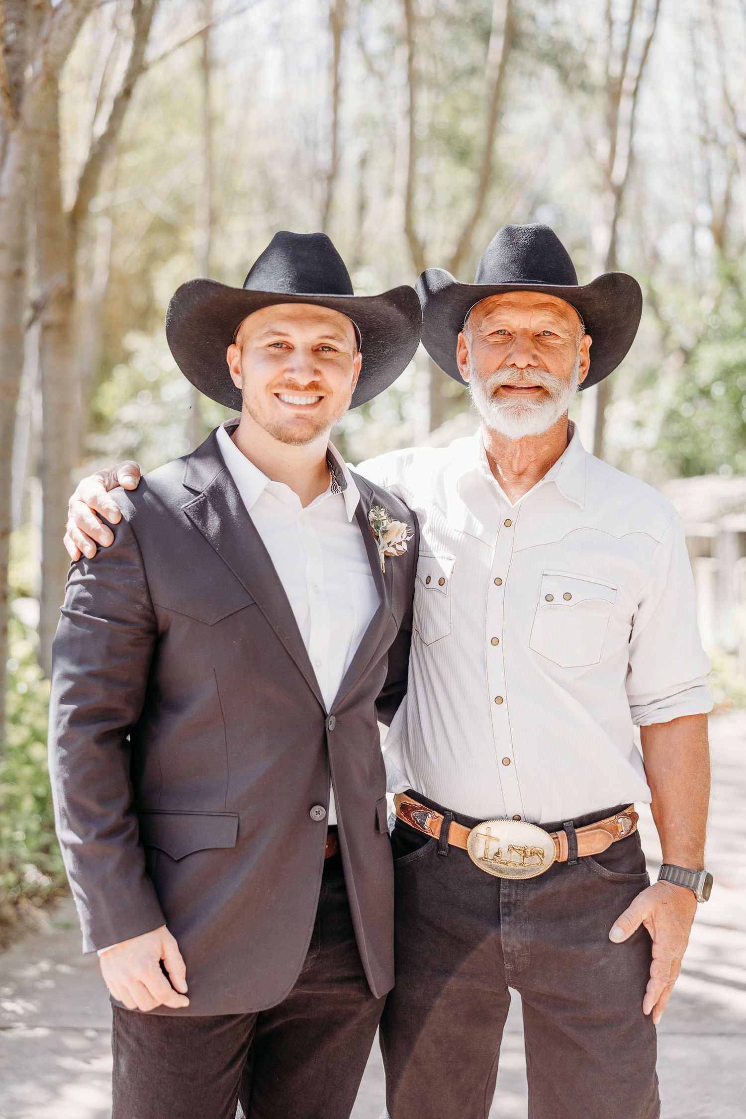 Two men in cowboy hats and western formal wear pose together at an outdoor wedding.