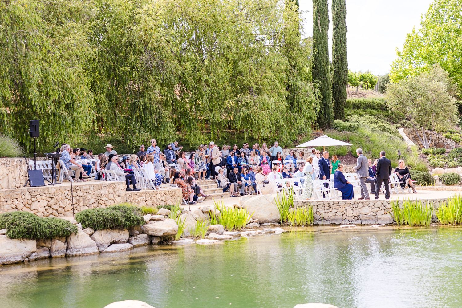 Outdoor wedding ceremony setup near a pond with guests gathered under willow trees.