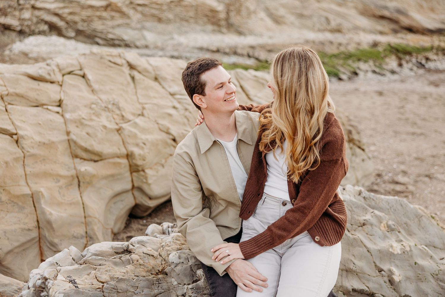 Two people embrace while sitting on rocky coastal cliffs at sunset.