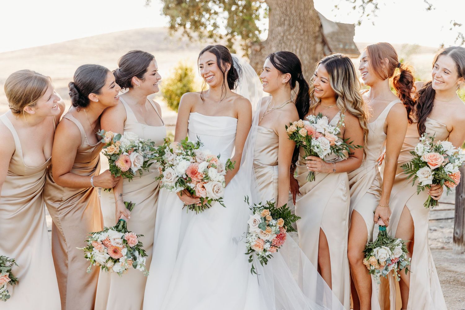Row of bridesmaids in champagne-colored dresses holding peach and white floral bouquets.