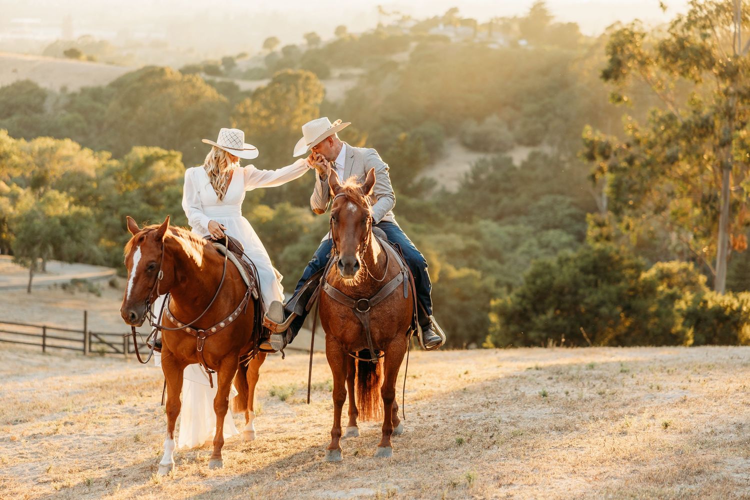 Two riders on brown horses interact while riding along a scenic hillside trail.