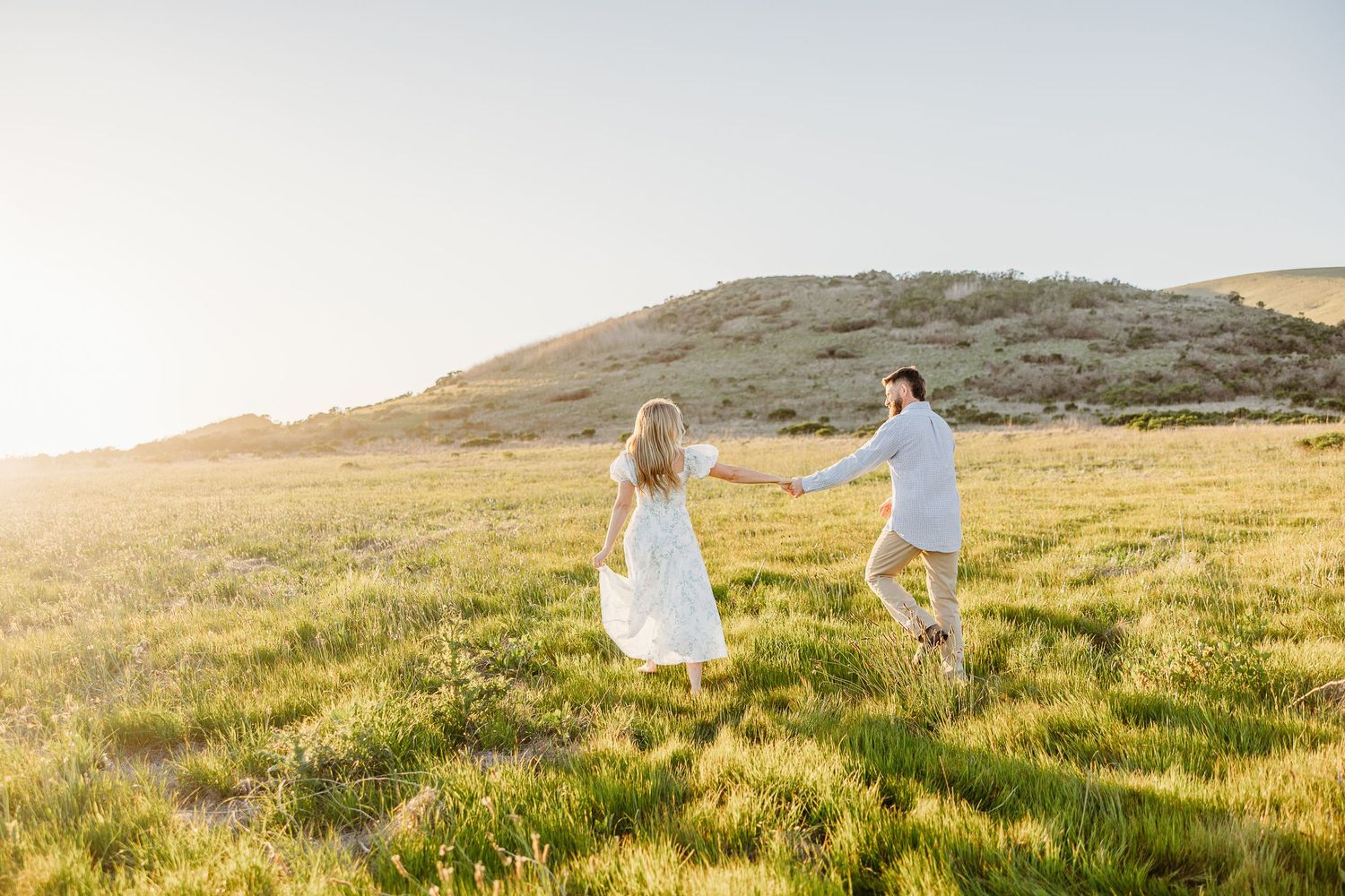 A couple in white outfits runs through a sunlit meadow on a grassy hillside at golden hour.