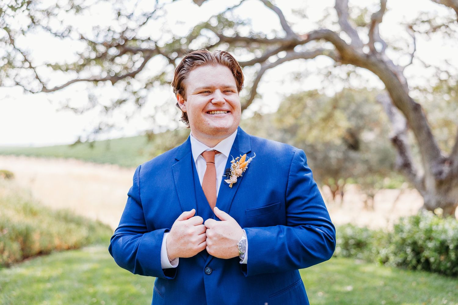 A joyful person in royal blue suit adjusts their tie while smiling outdoors.