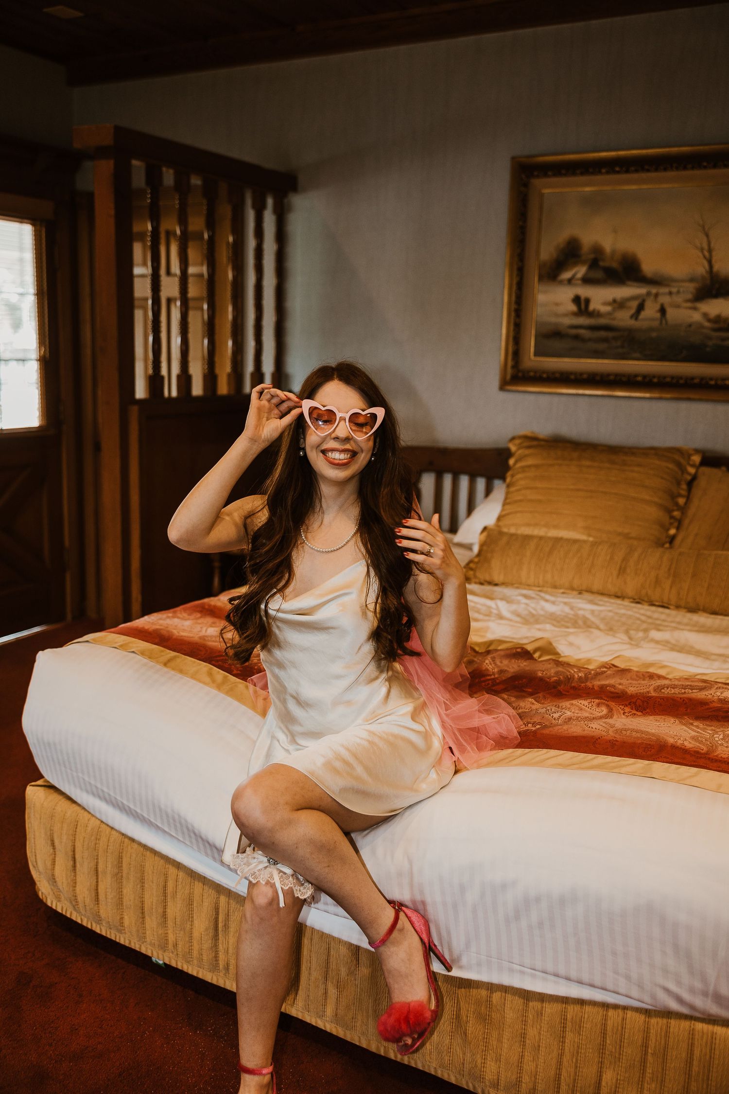 A person in a white dress sits on a bed wearing heart-shaped sunglasses in a warmly lit vintage-style bedroom.