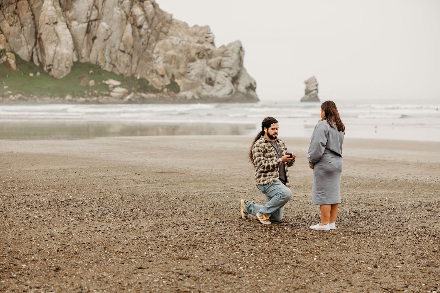 Two people share a special moment on a misty beach with dramatic coastal cliffs in the background.