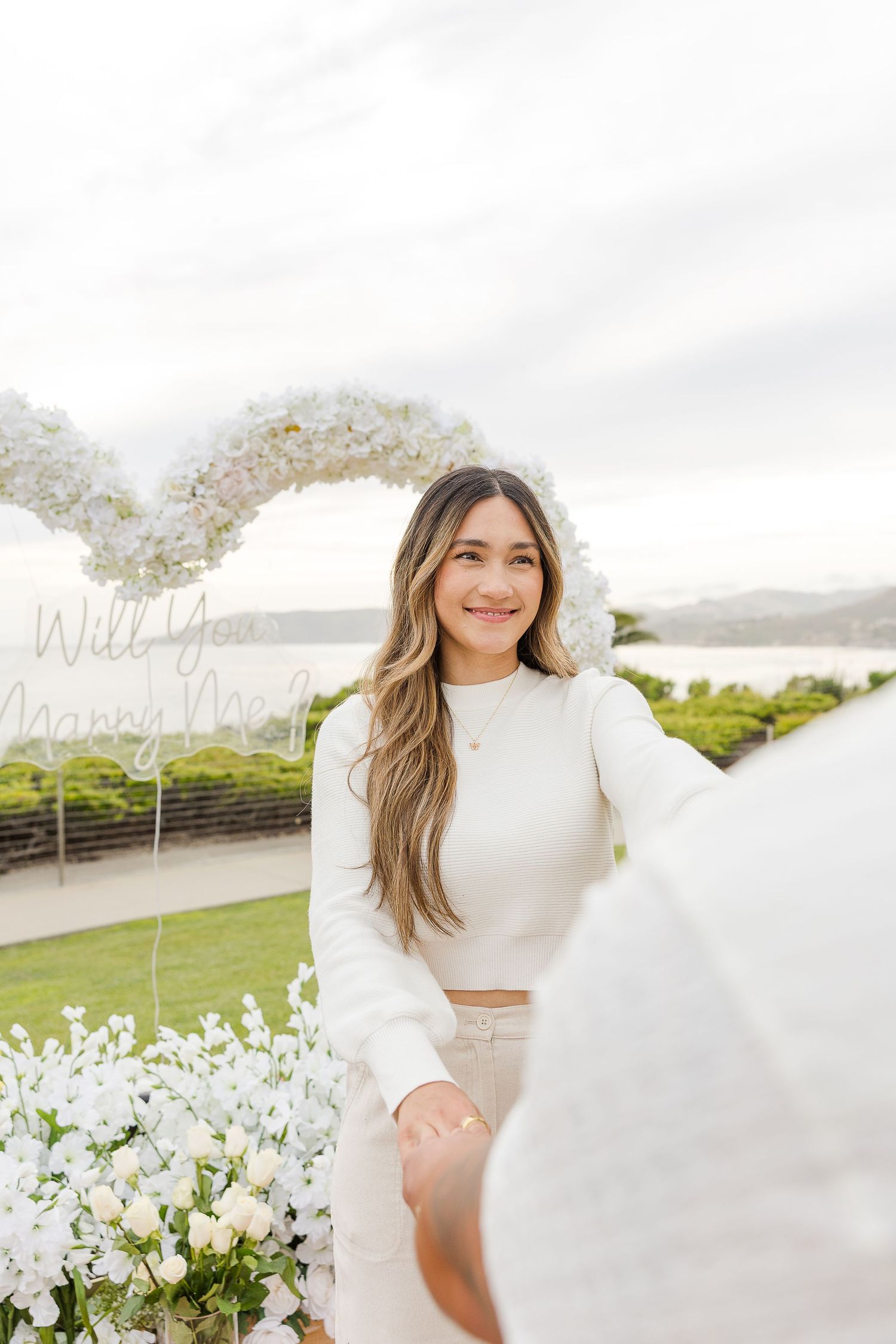 A romantic proposal scene unfolds against a coastal backdrop with white floral arrangements and natural scenery.