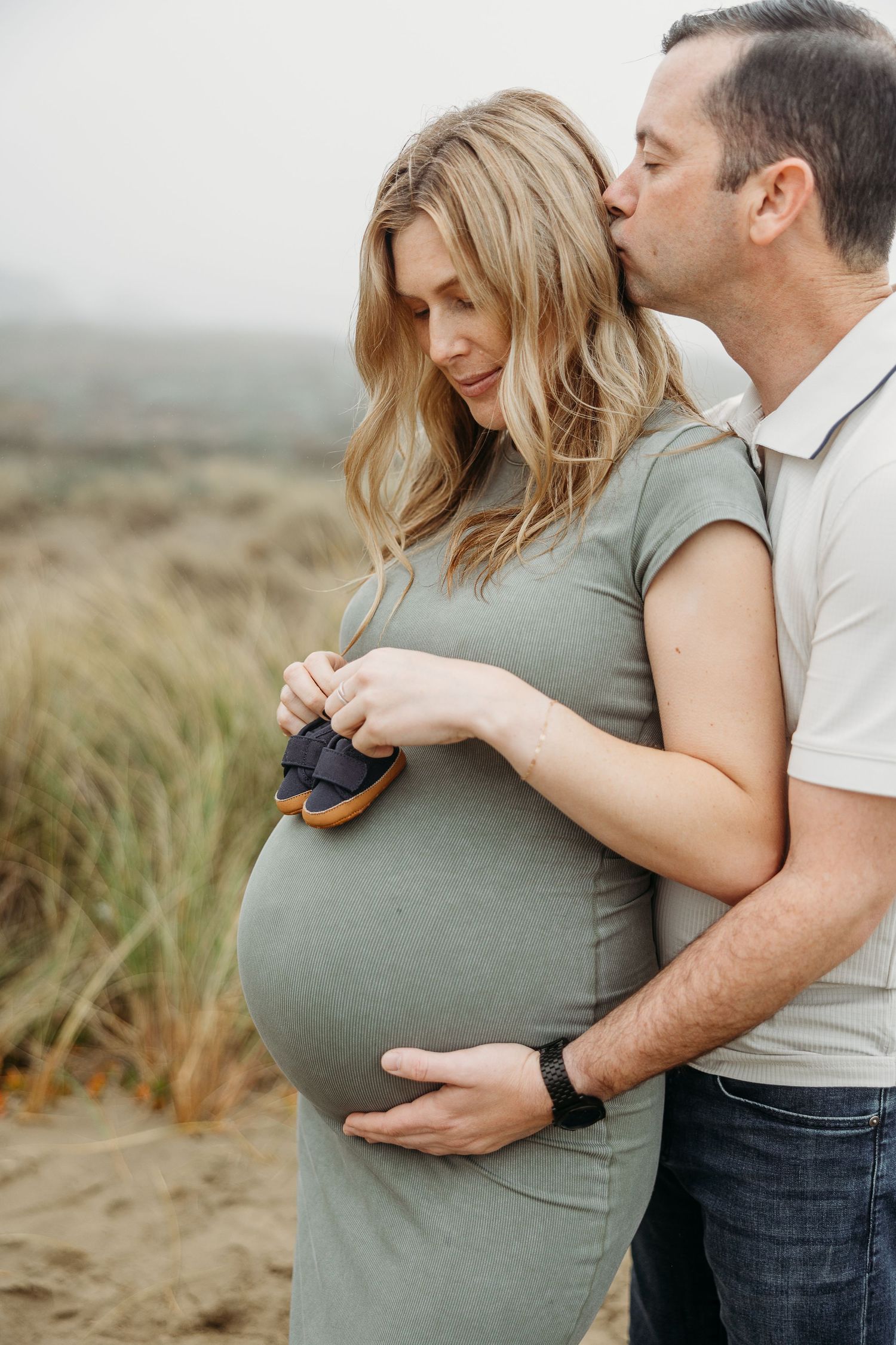 Intimate pregnancy photo shows gentle embrace and baby shoes against sage dress.