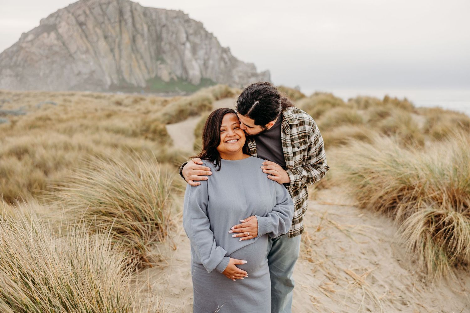 Intimate maternity photo of couple sharing tender moment against coastal landscape with mountain backdrop.