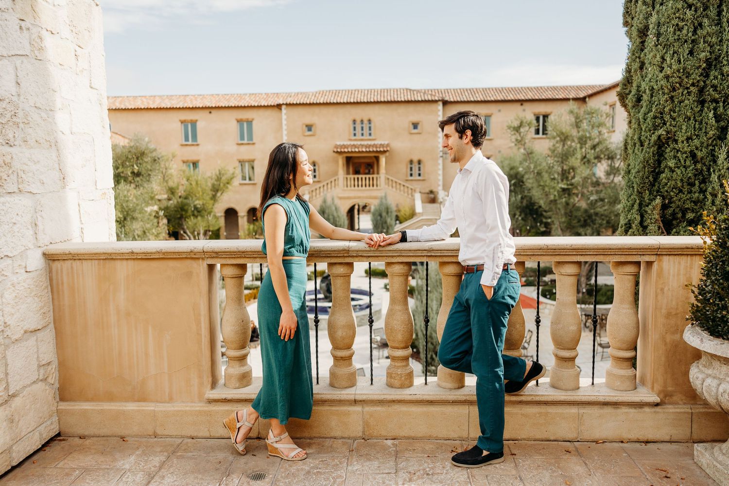 Two people stand on a Mediterranean stone balcony of a villa, wearing elegant summer attire.