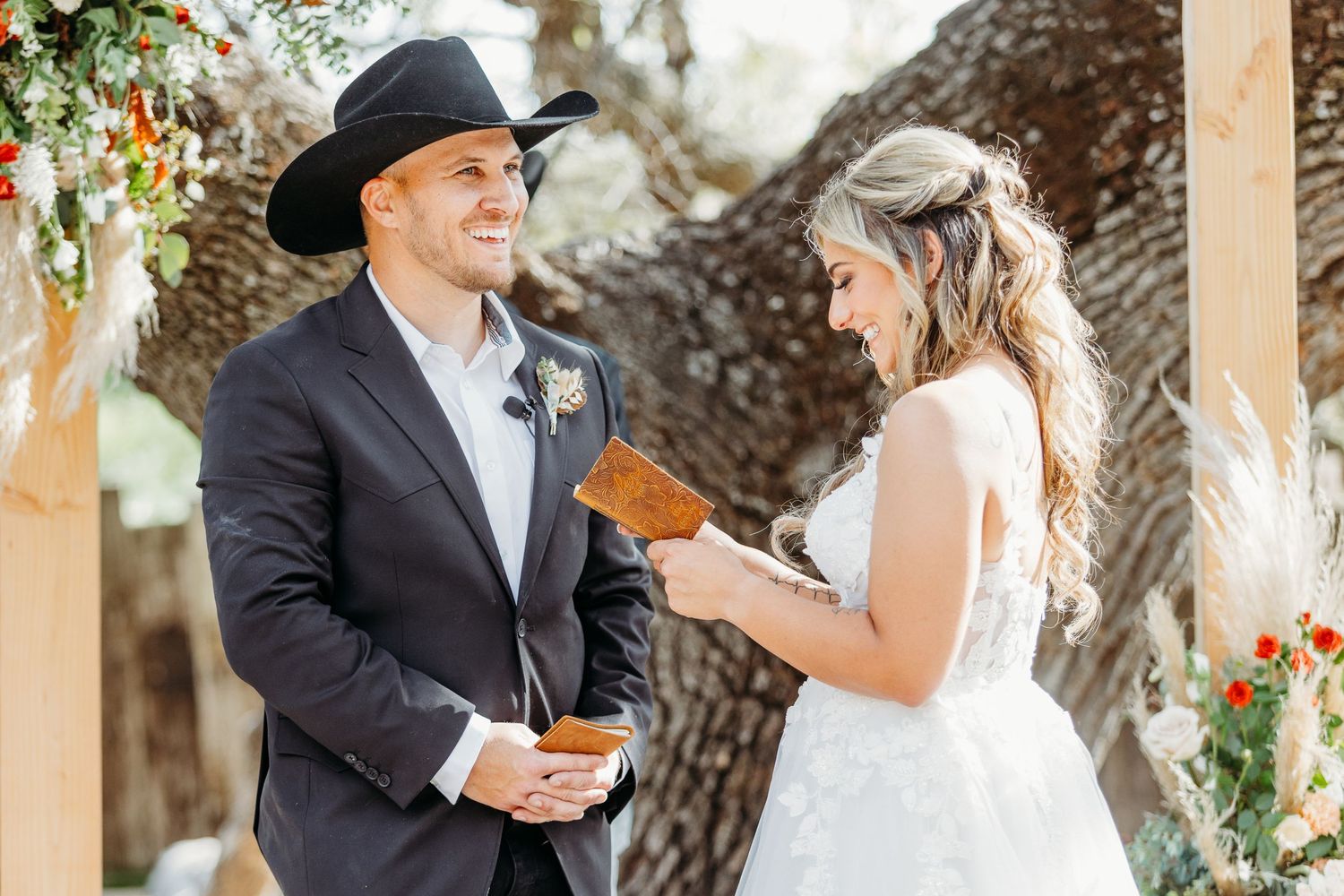 A couple exchanges vows at a rustic outdoor wedding ceremony adorned with floral decorations near a large tree.