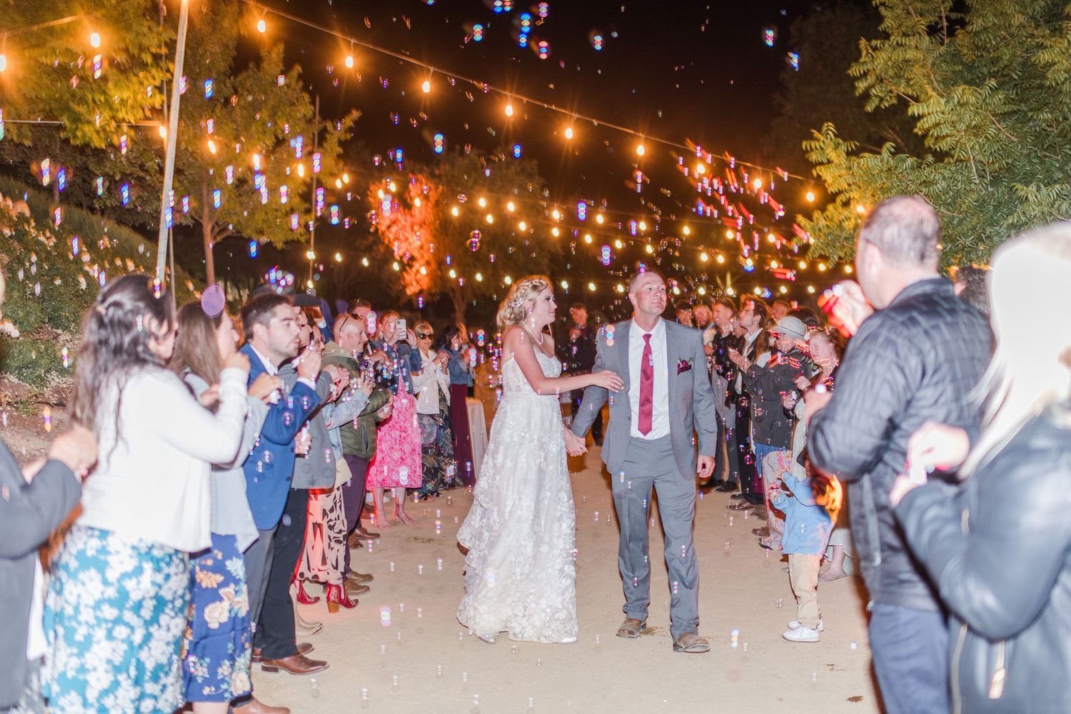 Wedding guests hold sparklers creating illuminated pathway for couple at nighttime celebration.