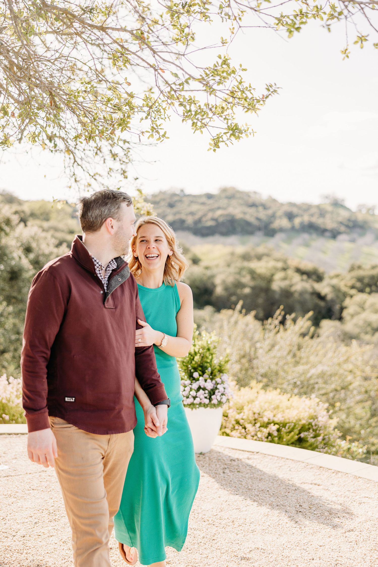 A couple shares a joyful moment outdoors on a path surrounded by trees and natural scenery.