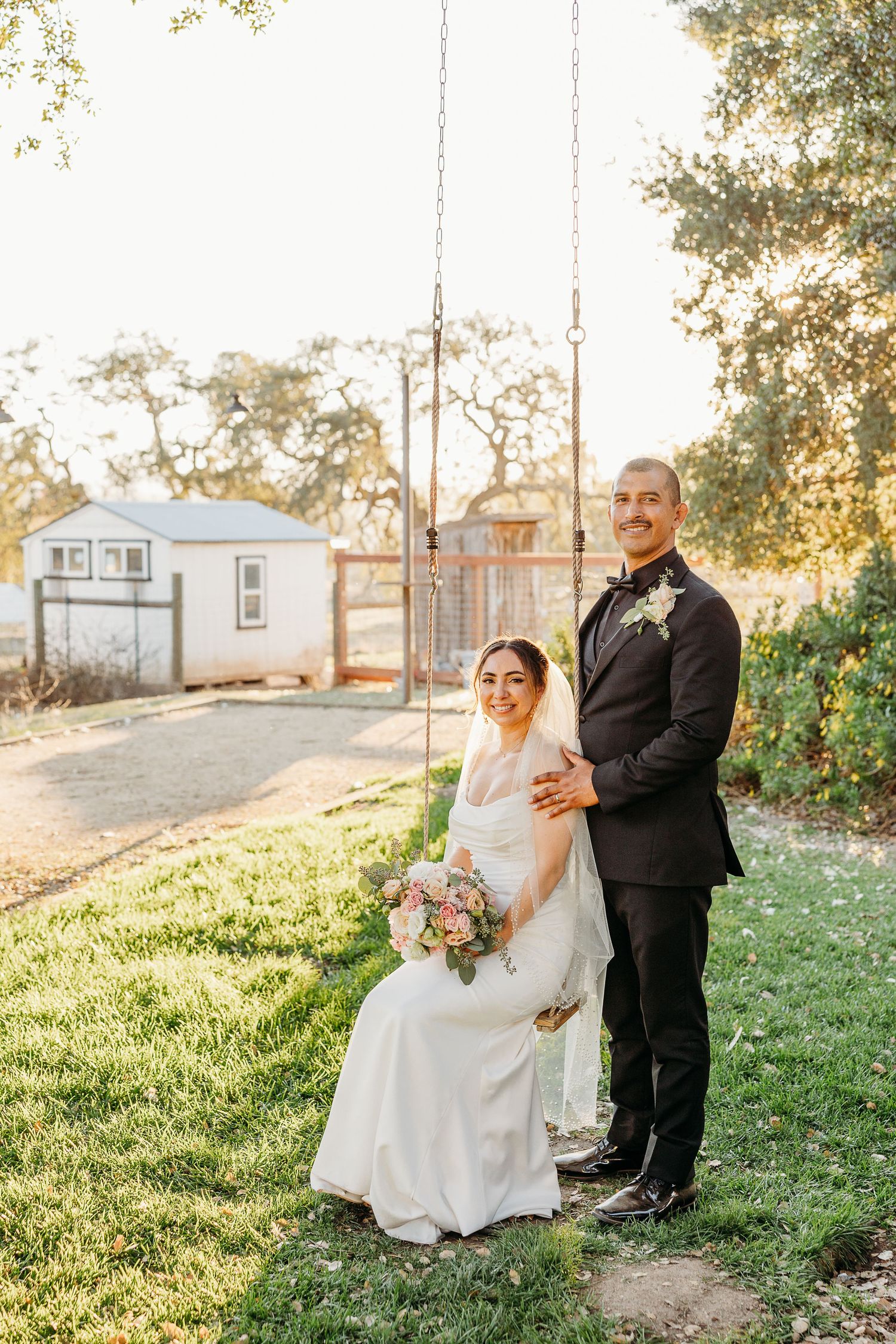 A romantic wedding photoshoot in a grassy field at golden hour with the couple sitting and laughing together.