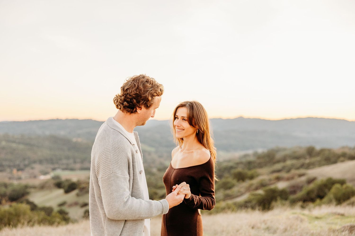 Couple embraces in romantic moment against backdrop of rolling hills during golden hour sunset.