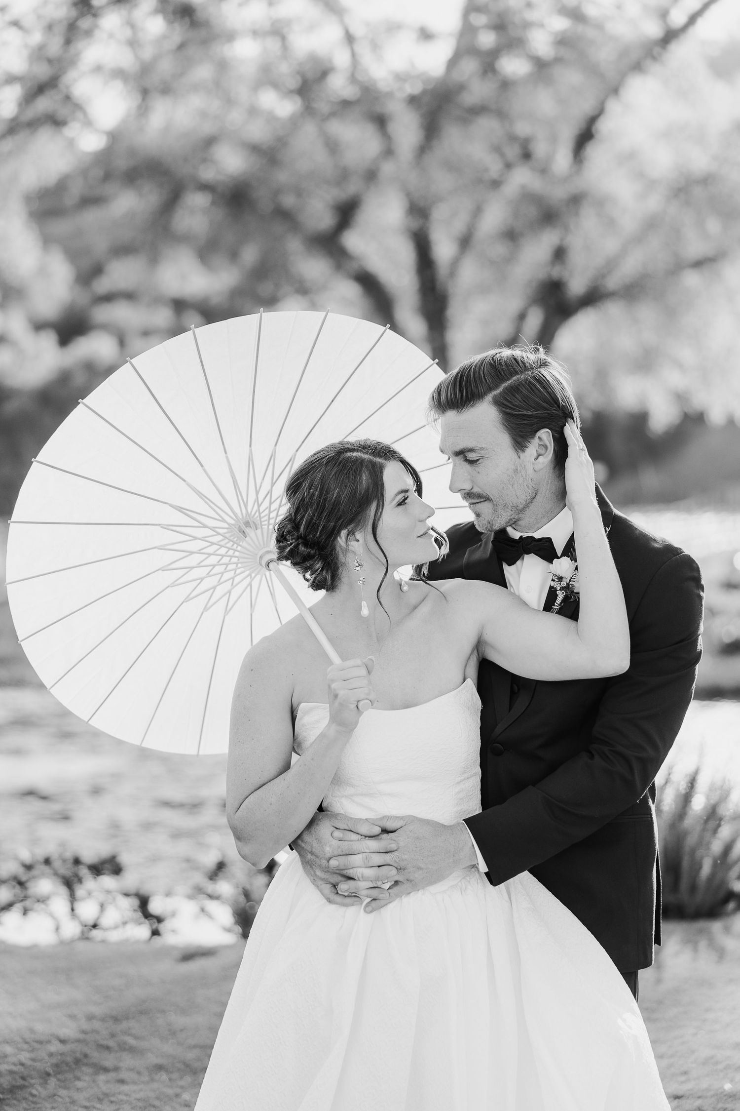A romantic black and white wedding photo featuring a couple sharing a tender moment under a white parasol outdoors.