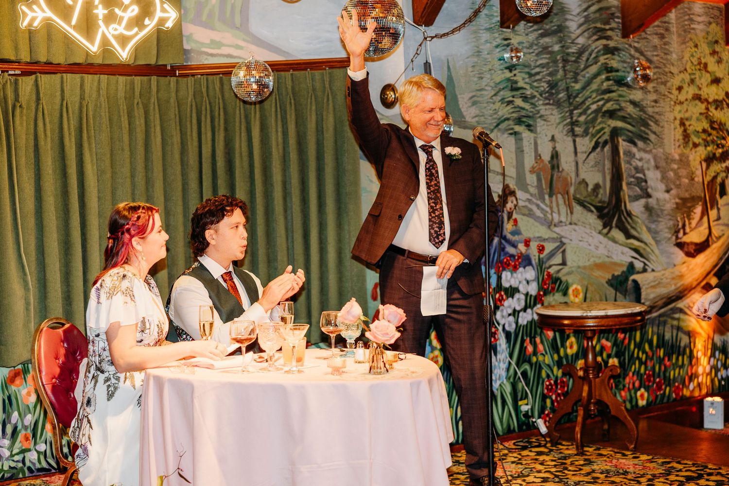 A festive wedding reception scene with guests seated at a decorated table while someone makes a toast.