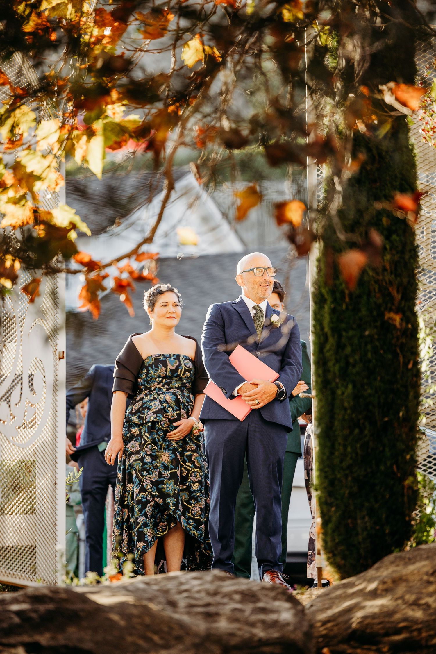 Wedding guests in formal attire mingling under trees with warm autumn lighting.