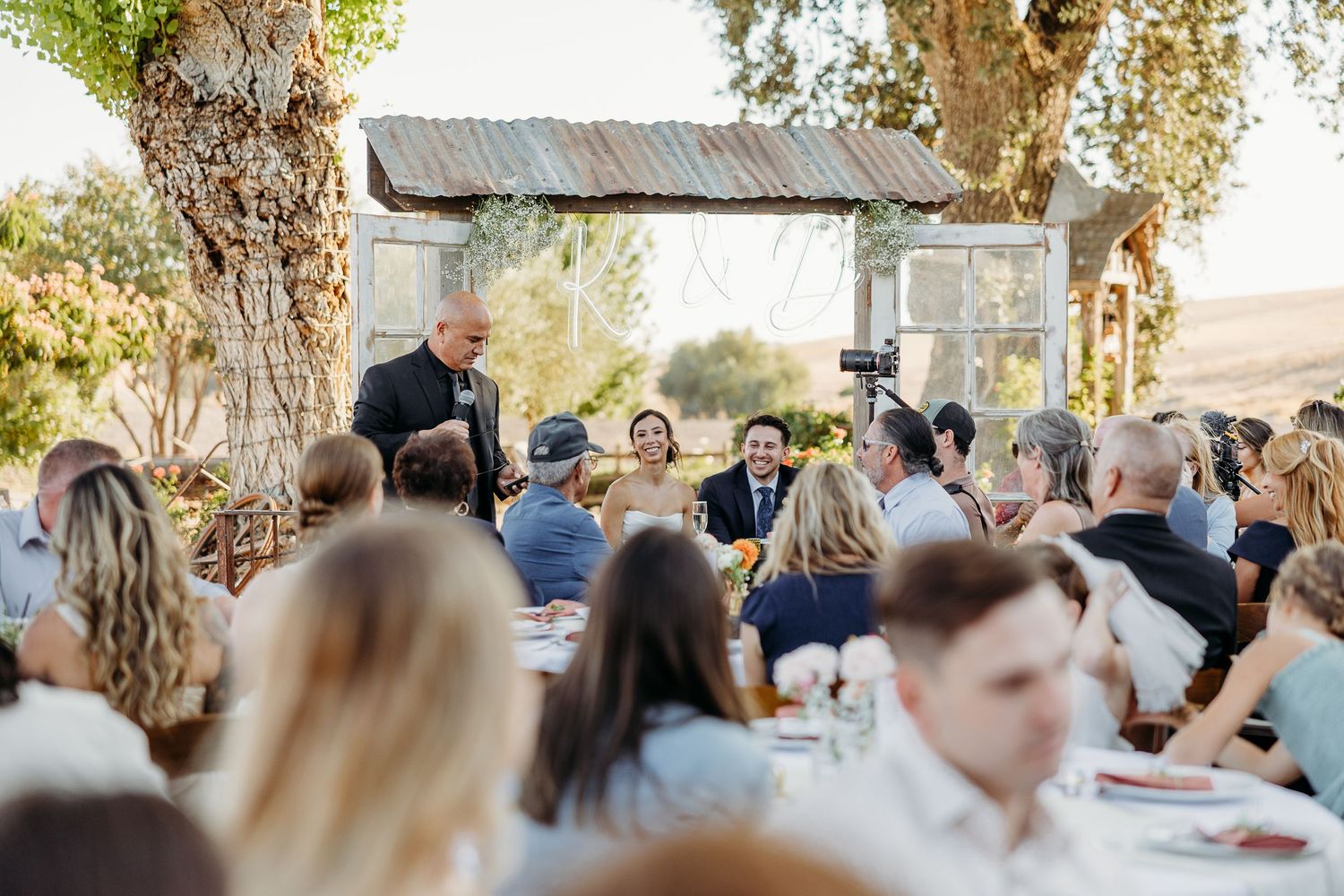 Outdoor wedding reception venue with guests seated under trees and string lights at dusk.