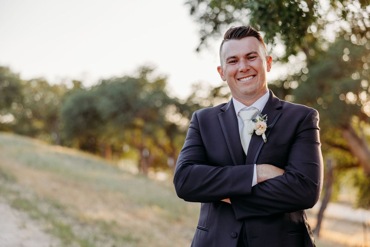 A groom smiling while wearing a classic black suit and tie outdoors during a wedding photoshoot.