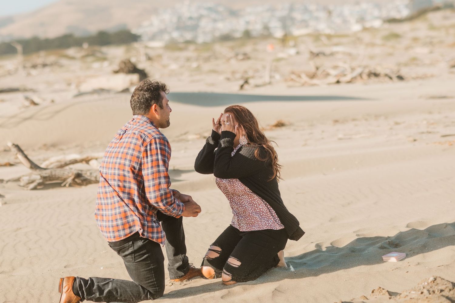 An emotional beachside proposal scene with both people kneeling in the sand.