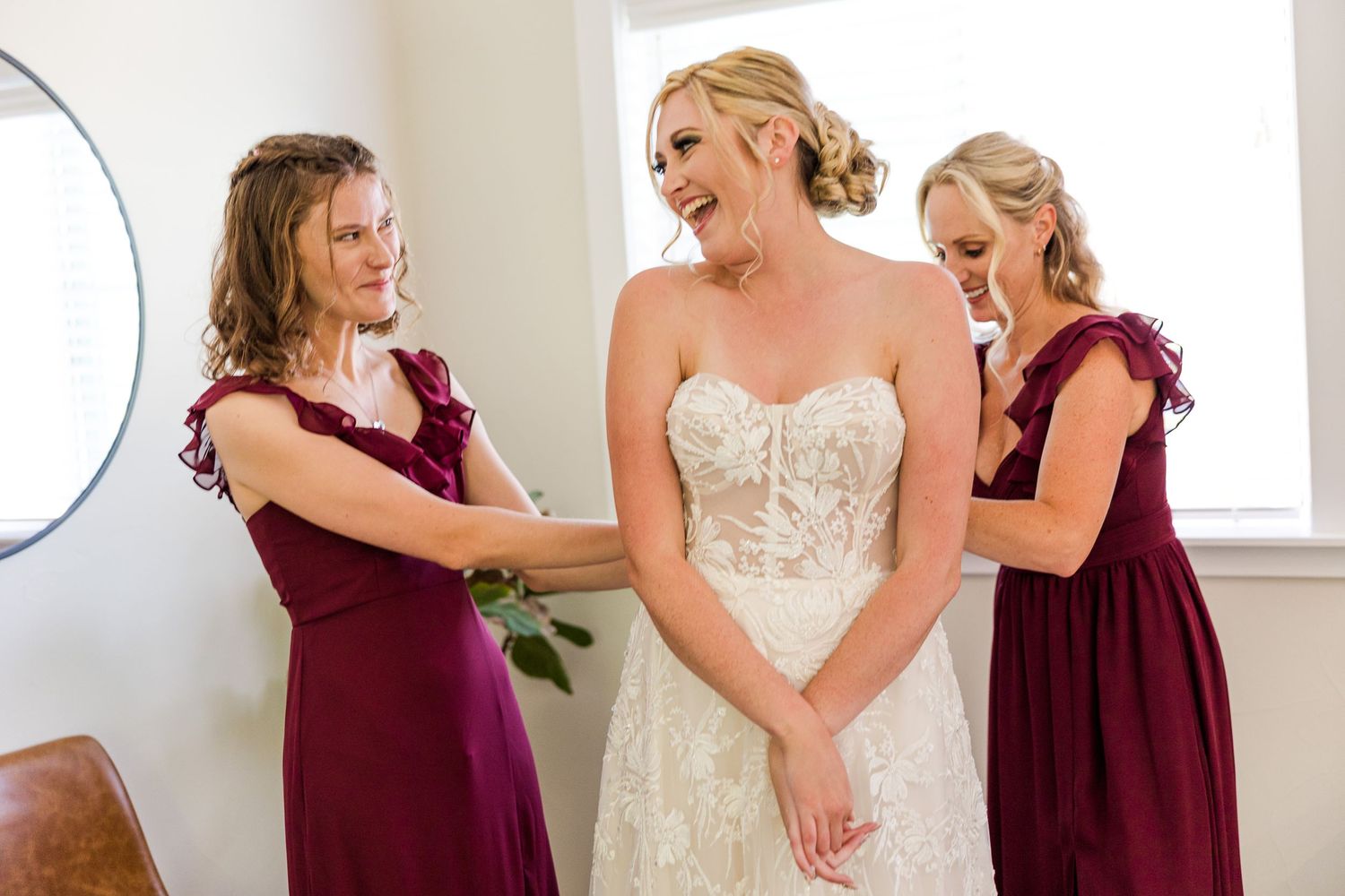 Bridesmaids in burgundy dresses help with final adjustments to a lace wedding gown in sunlit room.