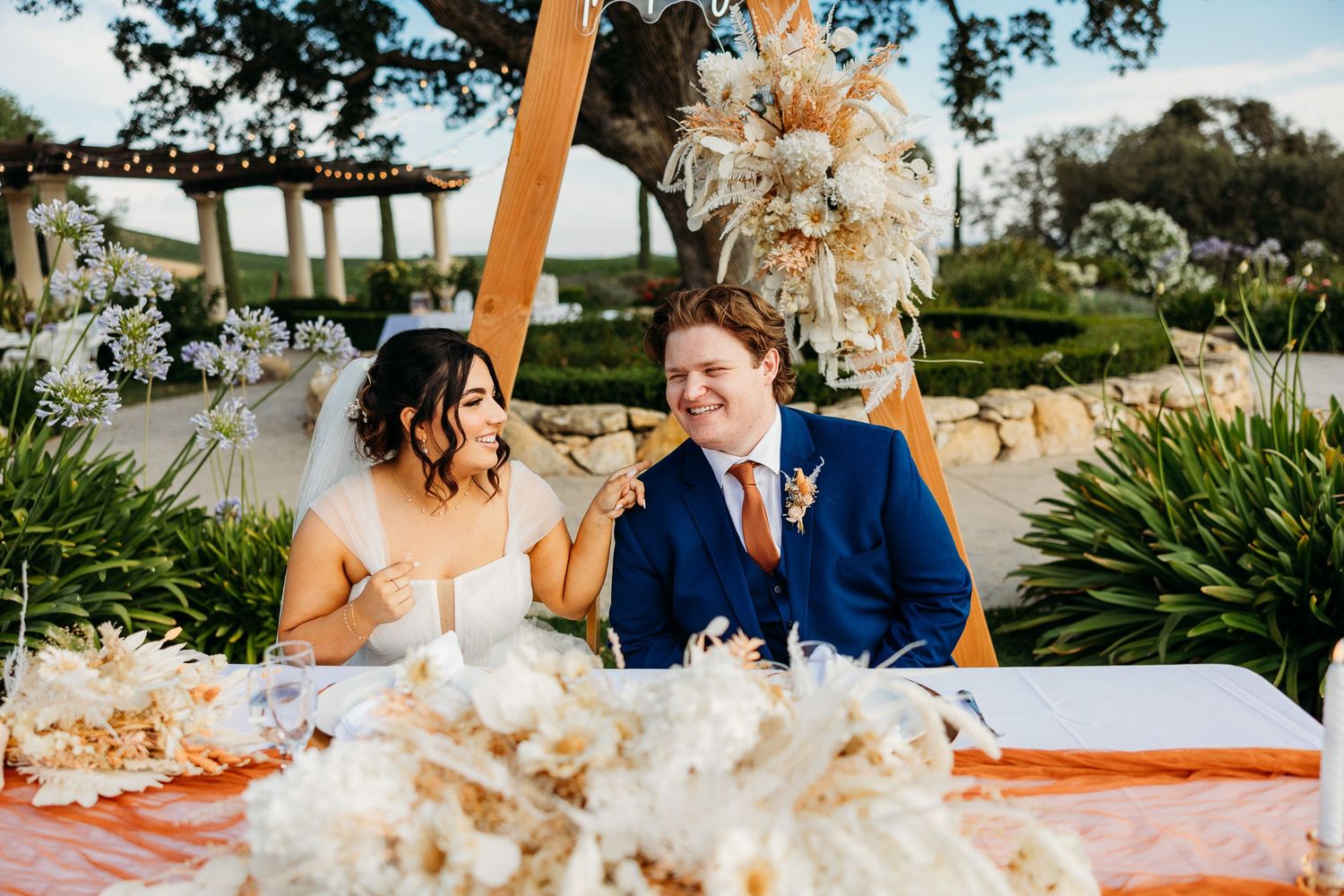 Couple seated at wedding table decorated with dried pampas grass arrangements in an outdoor garden setting.