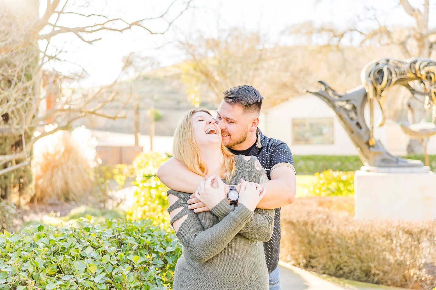 Sun-drenched portrait of couple embracing by garden hedges and bare trees.