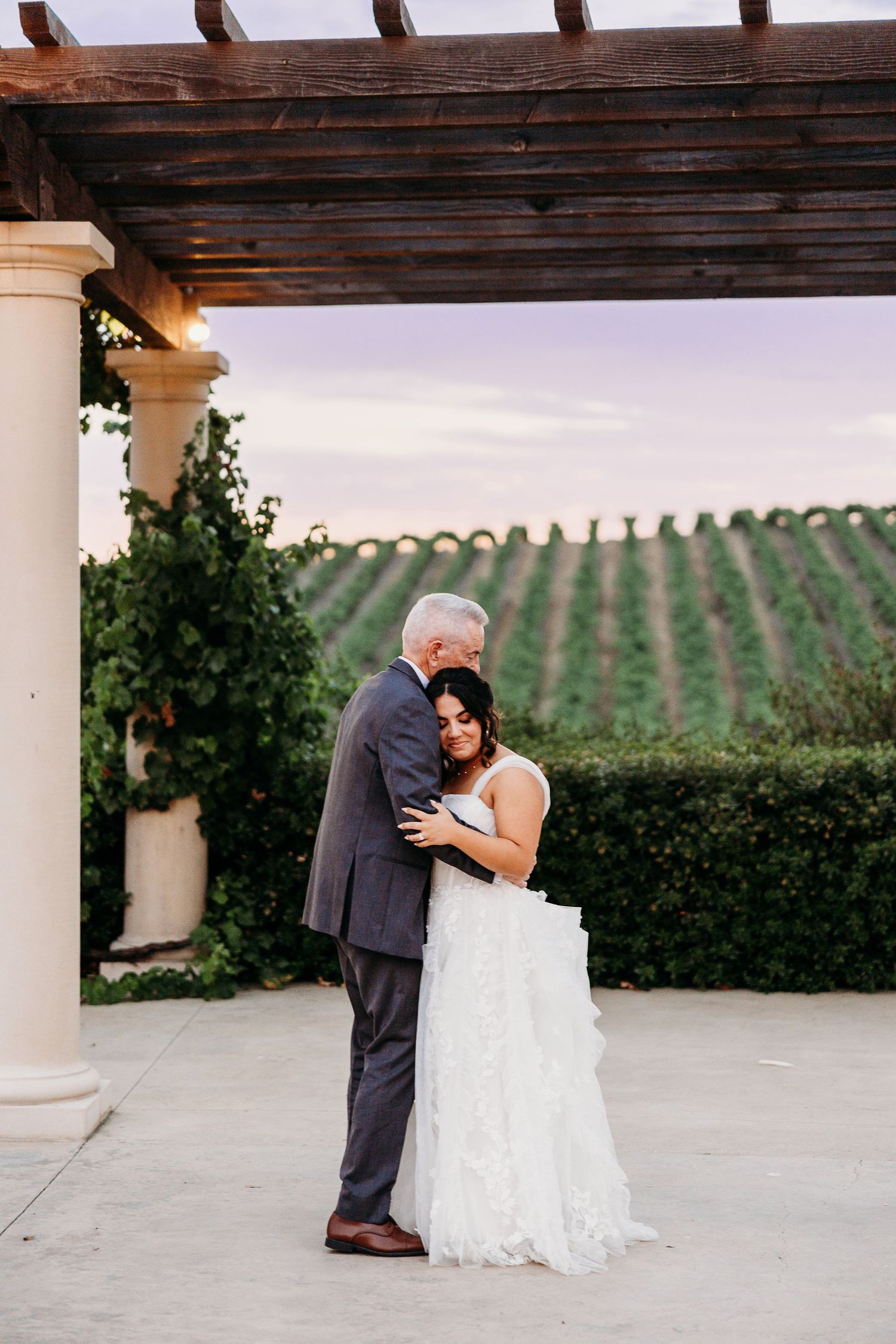 A tender wedding moment shared under a pergola with rolling vineyard hills in the background.