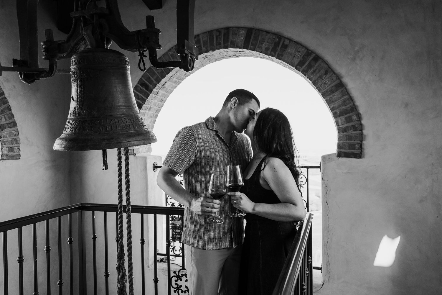A romantic black and white photo of a couple sharing an intimate moment beneath a bell tower arch.