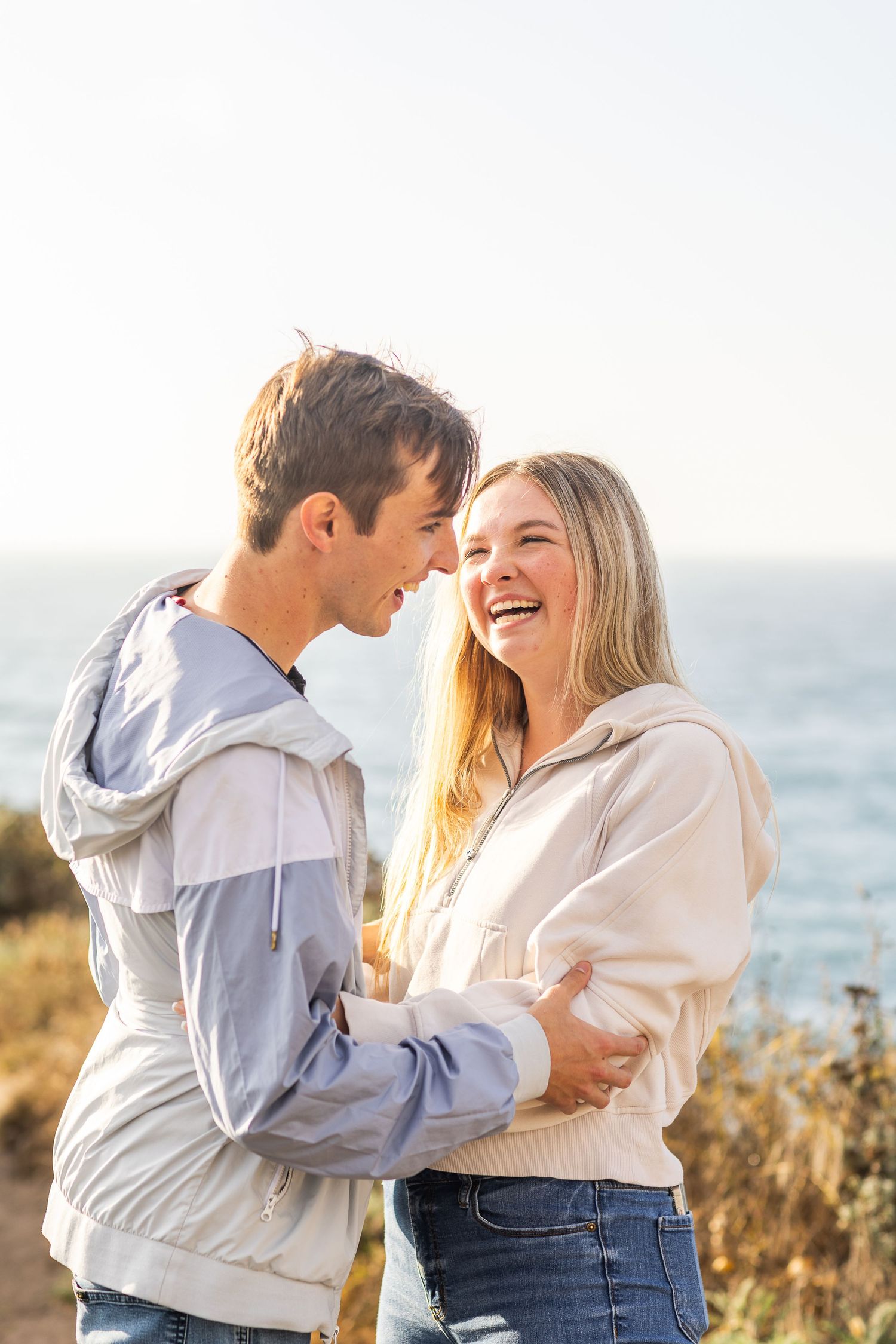 A joyful couple shares a laugh while embracing on a scenic coastal path.
