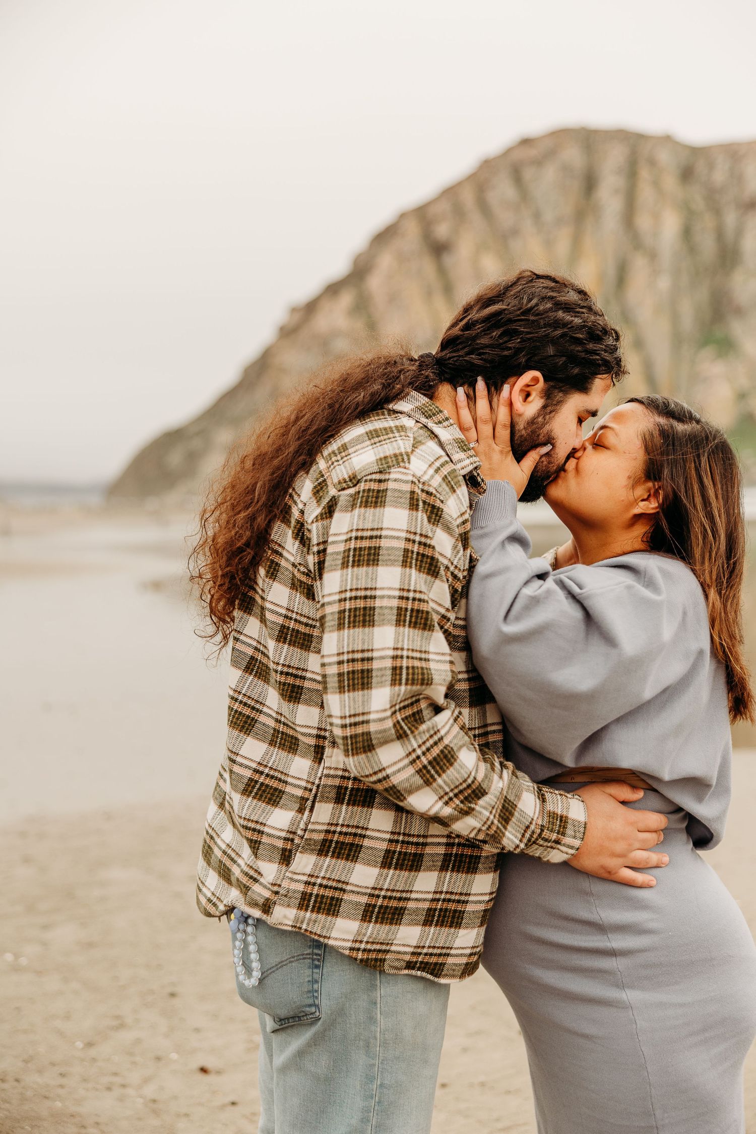 A romantic embrace between a couple on a beach with a large rock formation in the background.