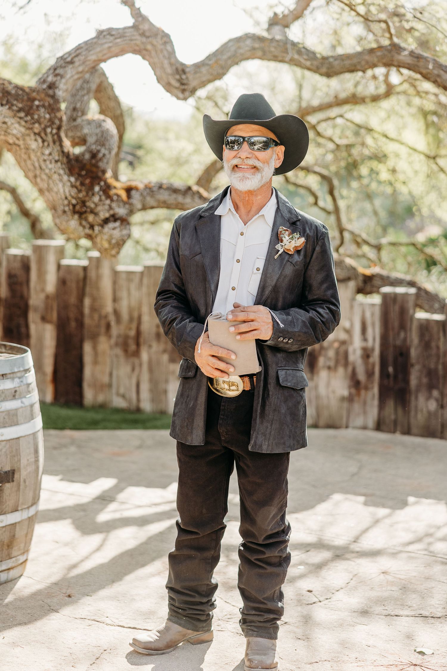 A distinguished gentleman in western attire with a cowboy hat and sunglasses poses at a rustic outdoor venue.