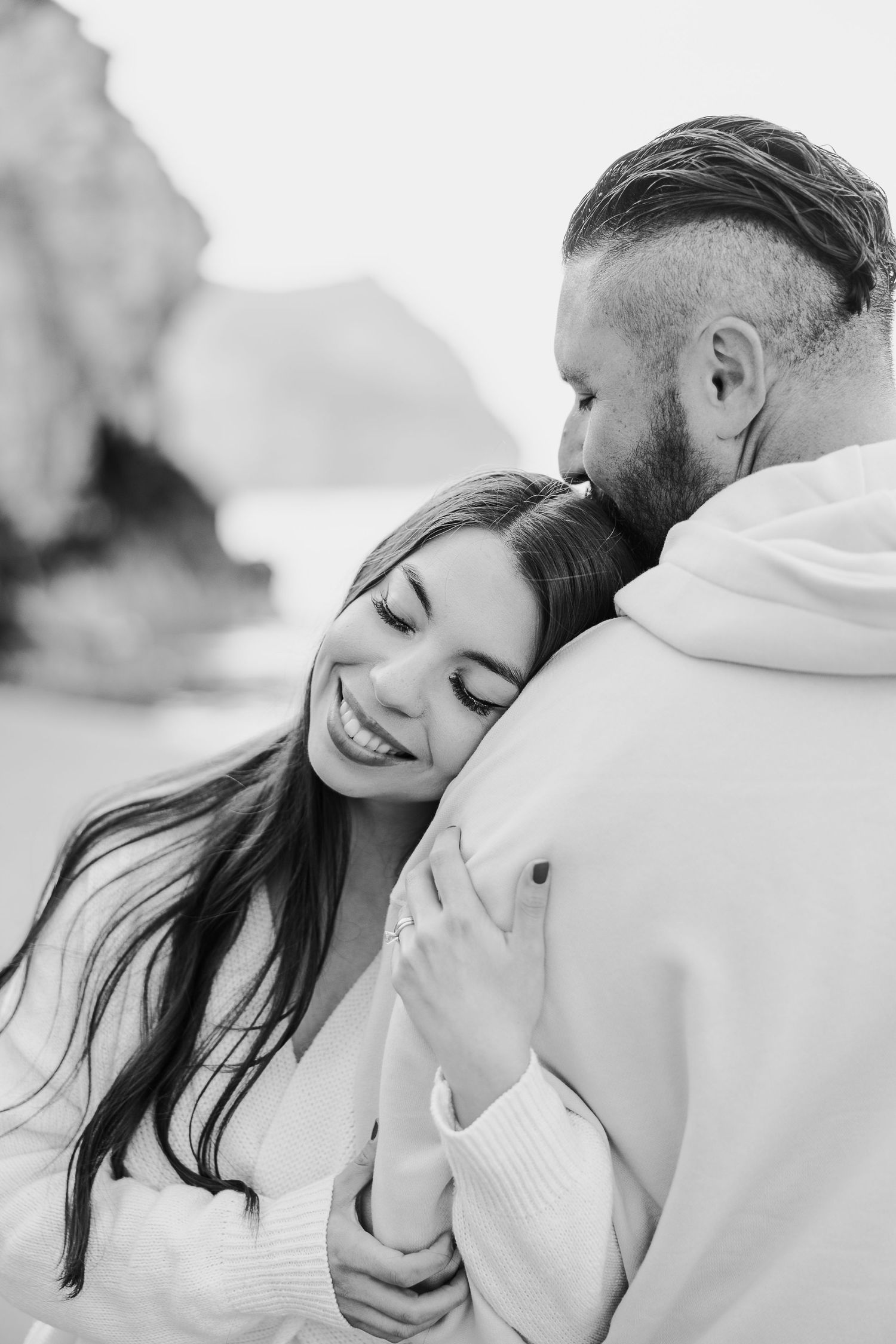 Black and white romantic photo of couple embracing outdoors with woman smiling and resting against partner's chest.