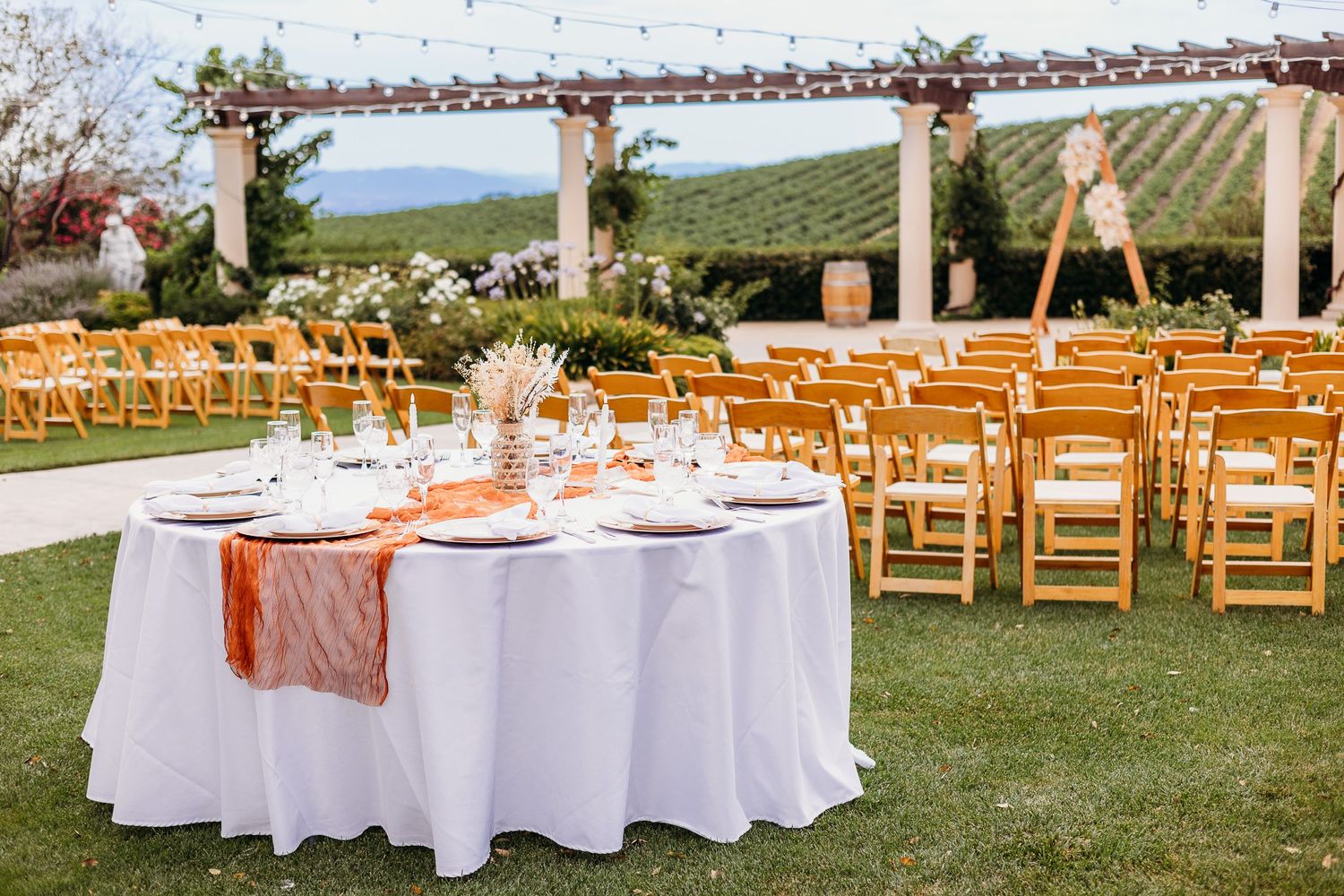 Wedding reception tables with white linens and orange accents set up under a rustic pergola.