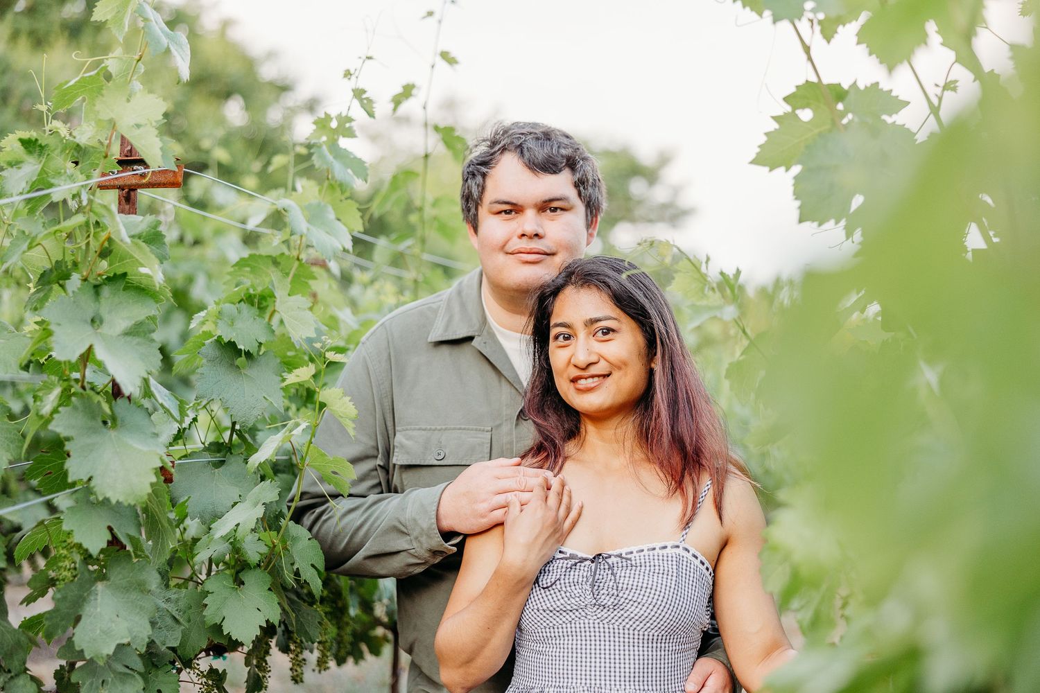 A couple smiles together while standing among grape vines in a sunny vineyard.
