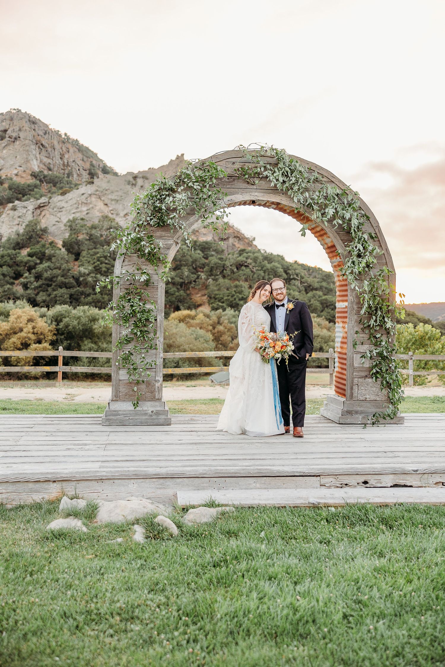 A romantic wedding scene under a vine-covered stone arch with mountain views and wooden deck platform.