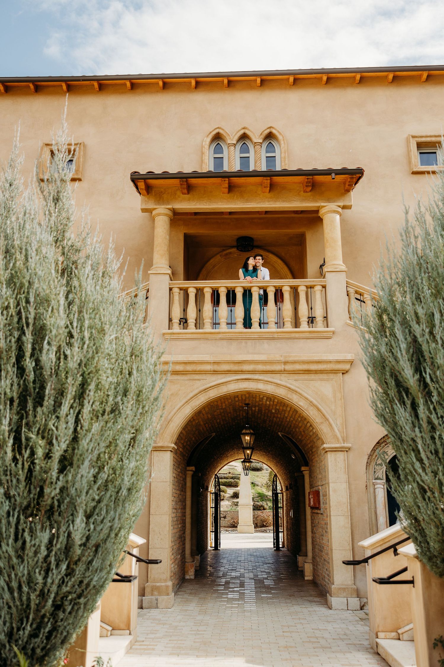 Mediterranean villa archway entrance with stone columns and decorative balcony, framed by tall cypress plants.