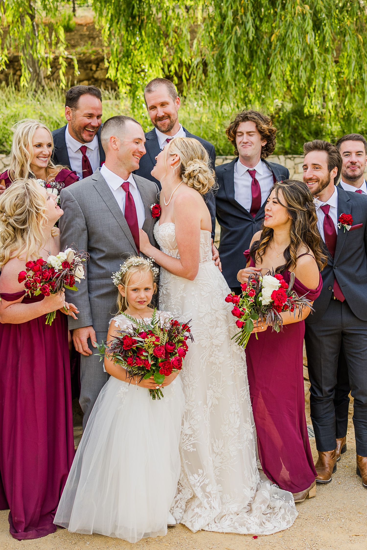 Wedding party in burgundy and gray attire gather for joyful group photo outdoors.