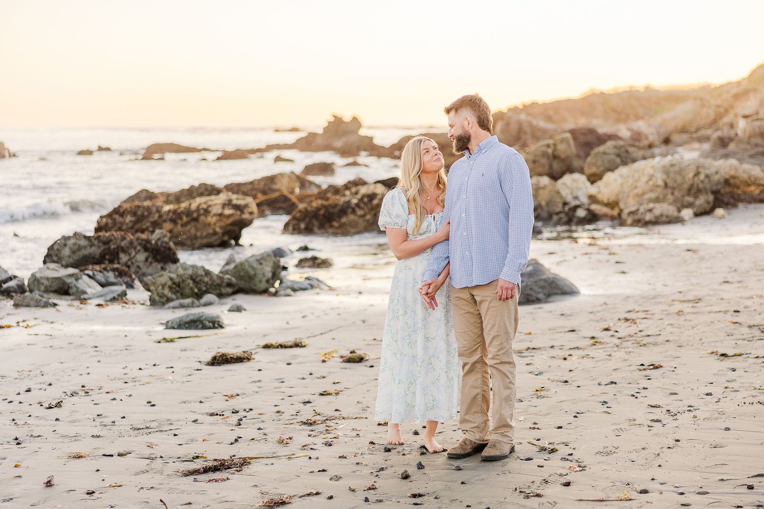 Couple strolling hand in hand along scenic beach at sunset with rocky cliffs.