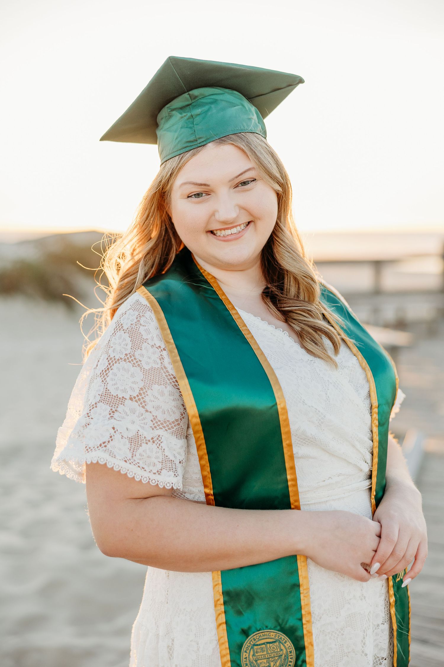 Graduate wearing green cap and gown with white dress poses on beach for graduation photos.