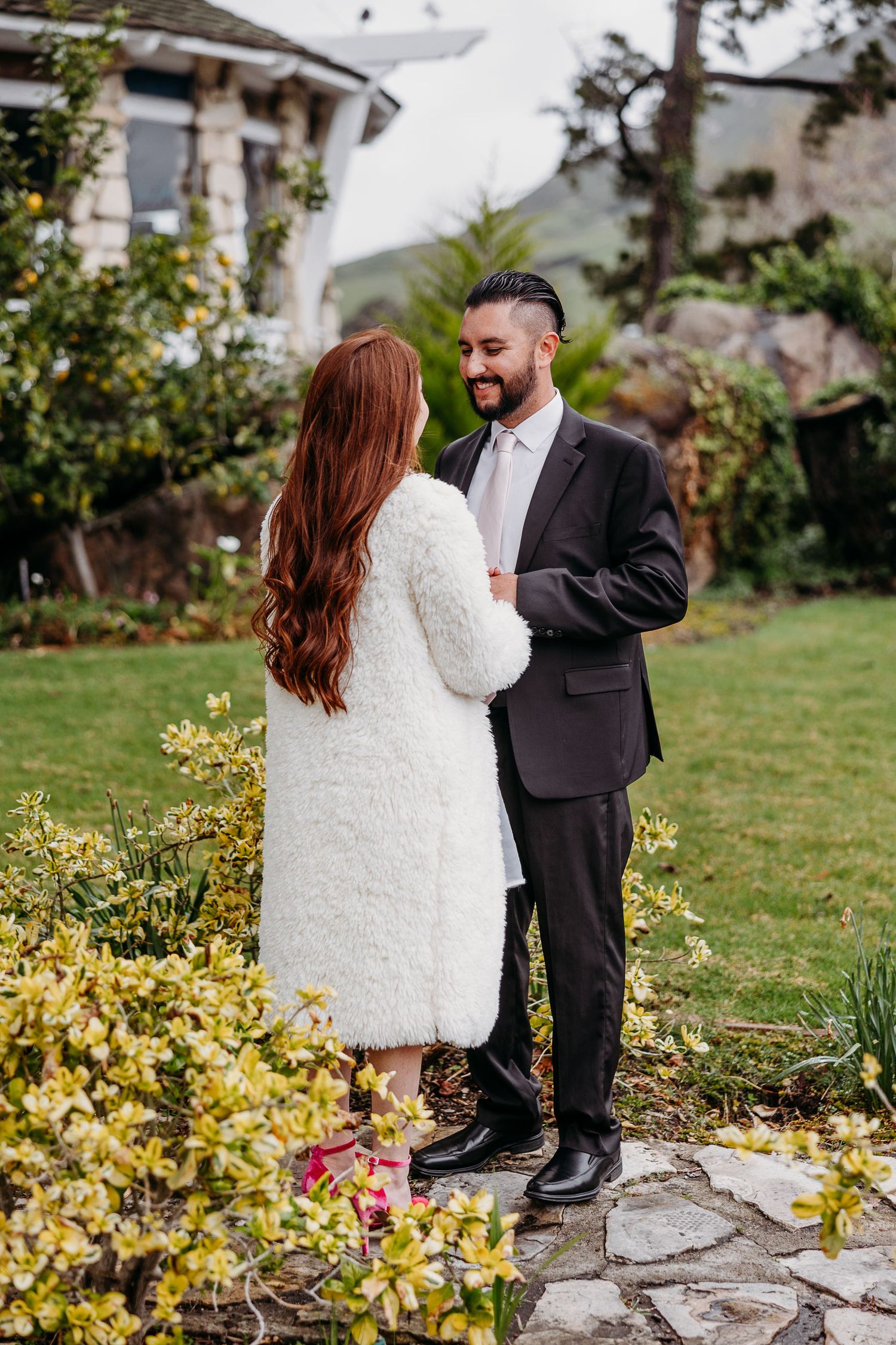 A couple stands together in a garden, with the person in white wearing a winter coat and the other in a dark suit.